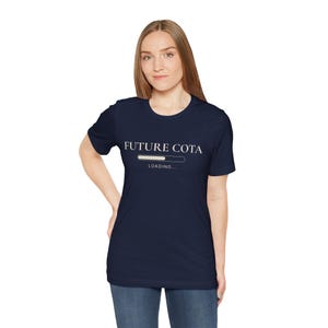 May include: A navy blue t-shirt with the text "FUTURE COTA" above a loading bar graphic. Below the bar, the word "LOADING..." is printed in a simple, sans-serif font. The shirt is a classic crew neck style.