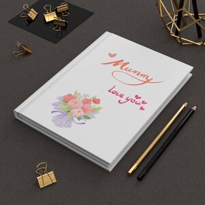 May include: White journal with the words "Mummy love you" in cursive script, accompanied by a bouquet of pink flowers. Gold and black pencils and gold binder clips are also visible.