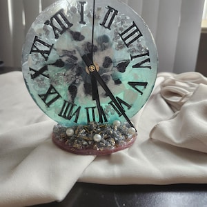 May include: A decorative clock with a clear, round face featuring black Roman numerals and black clock hands. The clock face has a teal and white design with embedded decorative elements. The base is a mix of grey, gold, and pink, adorned with pearls.