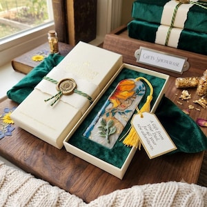 May include: A rectangular bookmark featuring a painted illustration of a person with a large yellow flower and green leaves, accented with gold flakes. It is presented in a cream-colored box with a wax seal and a yellow tassel with a gift tag.