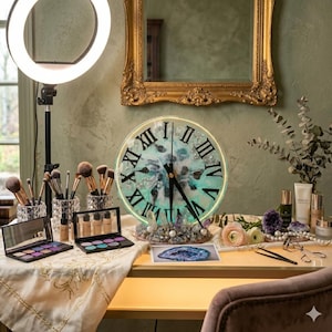 Könnte beinhalten: Ein Schminktisch-Arrangement mit einer dekorativen Uhr mit römischen Ziffern, Make-up-Pinseln und Lidschattenpaletten. Die Uhr hat ein türkis-silbernes Design. Ein Ringlicht und ein goldgerahmter Spiegel sind ebenfalls zu sehen.