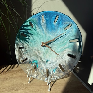 Puede incluir: Un reloj redondo con una esfera de resina transparente, con remolinos azules y turquesas, y manecillas de madera. El reloj tiene un soporte blanco y está decorado con pequeños adornos de color claro. El reloj tiene aproximadamente 15 cm de diámetro.