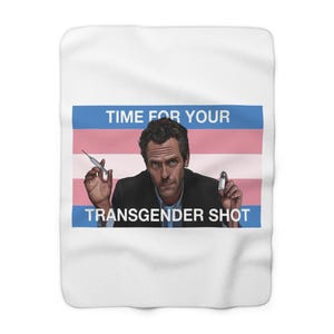 Könnte beinhalten: Weiße Fleece-Decke mit einem Bild eines Mannes, der eine Spritze und ein kleines Fläschchen hält. Das Bild enthält den Text "TIME FOR YOUR" oben und "TRANSGENDER SHOT" unten, vor der Transgender-Pride-Flagge.