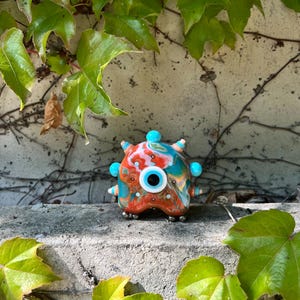 Op de afbeelding: Een kleurrijke glazen sculptuur die lijkt op een klein, grillig wezen. Het heeft een mix van oranje, blauwe en witte tinten met een centraal oogachtig ontwerp. De sculptuur heeft verschillende blauwe bolvormige uitsteeksels en staat op een betonnen richel, omringd door groene bladeren.