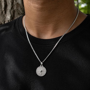 May include: Silver compass pendant necklace. The round pendant features a detailed compass design with a black centre. The necklace is a silver chain, and the pendant hangs from the chain. The person is wearing a black t-shirt.
