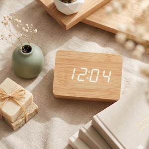 Puede incluir: Un reloj digital de madera rectangular que muestra las 12:04. El reloj tiene una esfera de color claro y está colocado sobre una superficie neutra. Otros elementos incluyen un pequeño jarrón verde con flores, jabones atados con hilo y una suculenta en una maceta blanca.