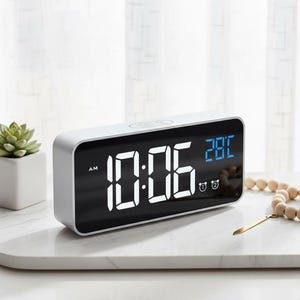 May include: A white digital alarm clock displaying 10:06 AM and 28°C. The clock has a rectangular shape with rounded corners and a black screen. Two alarm clock icons are visible. A small succulent plant in a white pot is on the left.