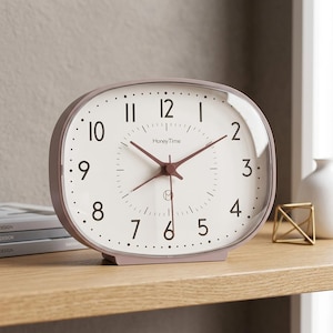 May include: An oval-shaped analog table clock with a dusty rose frame and a white face. The clock features dark brown Arabic numerals, matching dark brown hour, minute, and second hands, and the text HoneyTime printed in the center of the dial.