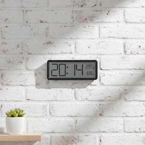 Puede incluir: Un reloj digital negro que muestra las 20:14, con lecturas de temperatura y humedad. El reloj está montado en una pared de ladrillos blancos. La temperatura es de 29,5 grados Celsius y la humedad es del 68%.