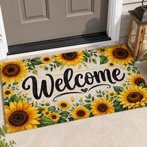 Può includere: Un zerbino rettangolare con la scritta "Welcome" in nero, circondato da vivaci girasoli gialli e foglie verdi su uno sfondo chiaro effetto legno. Il tappeto è posto davanti a una porta grigia.