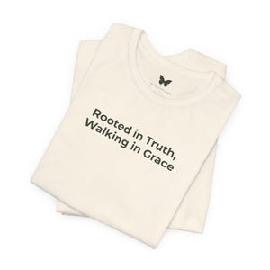 Op de afbeelding: Twee crèmekleurige T-shirts, opgevouwen en op elkaar gestapeld, met de tekst "Rooted in Truth, Walking in Grace" in zwarte letters. Een klein vlinderlogo is zichtbaar bij de halslijn. De shirts lijken van zacht materiaal te zijn.