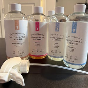 May include: Four clear bottles of multi-purpose cleaner with white labels and white caps. The labels feature the text "Multi-Purpose Cleaner" and different scent names: White Ginger Peach, Pinktastic, Fresh Air Care+, and Island Breeze. A white spray nozzle is in the foreground.