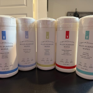 May include: Five white cylindrical containers of multi-purpose wipes. Each container has a different colored band at the bottom and a label with a scent name: Island Breeze, Fresh Air Care, Honeydew Breeze, Radiant Raspberry Air Care, and Coconut Water Lily.