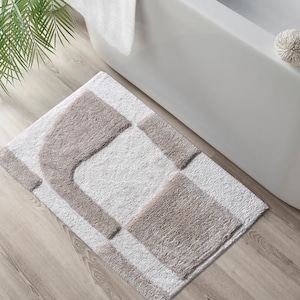May include: A rectangular bath mat featuring a high-pile, tufted texture in a two-tone color scheme. The design consists of a white base with abstract, geometric shapes in a soft taupe color, including a curved arch-like form and a square block.