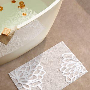 May include: A rectangular bath mat in a light beige color featuring a raised white floral pattern on both ends. The mat has a textured, shaggy pile surface and is positioned on the floor next to a curved, off-white bathtub filled with water.