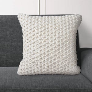 May include: A square, cream-coloured decorative cushion with a textured, woven pattern. The cushion is placed on a grey sofa, offering a neutral colour palette for home decor.