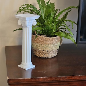 May include: A white, miniature Corinthian column with fluted sides and ornate capital. The column is displayed on a dark wood surface, with a large green plant in a woven basket in the background. The column is approximately 6 inches tall.