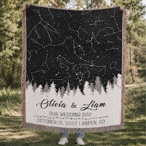Könnte beinhalten: Eine gewebte Wohndecke mit einem schwarzen Nachthimmel voller weißer Sternbilder über einer weißen Waldsilhouette. Der Text lautet Olivia & Liam, OUR WEDDING DAY, und OCTOBER 15, 2022 | ASPEN, CO, ergänzt durch eine dekorative Linie aus kleinen Kreisen.
