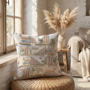 May include: A cushion with a watercolour design of a bookshelf filled with books, plants, and butterflies. The word "Matilda" is written on the middle shelf. The cushion is on a woven wicker ottoman near a window.