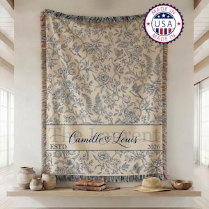 Könnte beinhalten: Eine gewebte Wohndecke mit cremefarbenem Hintergrund und einem blauen botanischen Muster aus Blumen, Farnen und Vögeln. Ein horizontaler Streifen zeigt den Text "Camille & Louis" mit "ESTD" und "2026" an den Seiten. Die Decke hat gefranste Kanten.