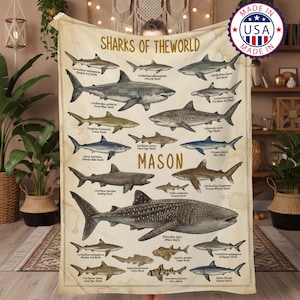 Puede incluir: Una manta beige con ilustraciones de varias especies de tiburones, con sus nombres científicos. La manta tiene el texto "SHARKS OF THE WORLD" y el nombre "MASON" impresos. Una pegatina "MADE IN USA" es visible.