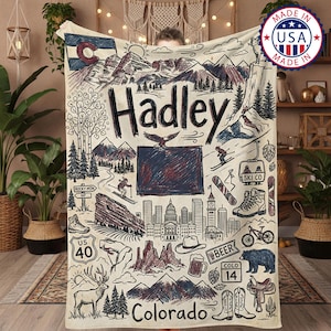 Puede incluir: Una manta color crema con una ilustración dibujada a mano de los puntos de referencia de Colorado, incluyendo montañas, un paisaje urbano, y los textos "Hadley" y "Colorado". El diseño incorpora una bandera estatal, un esquiador y otros iconos. La manta está marcada "Made in USA".