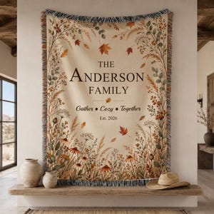 Op de afbeelding: Een geweven plaid met een crèmekleurige achtergrond en een botanische rand van herfstbladeren en wilde bloemen. De tekst luidt THE ANDERSON FAMILY, Gather • Cozy • Together en Est. 2026. De deken heeft franjes aan de randen en warme tinten.