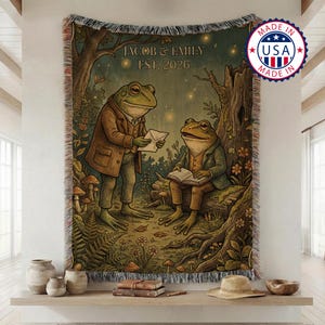 May include: Woven tapestry throw blanket featuring a whimsical illustration of two anthropomorphic frogs in a forest setting. One frog holds a letter, the other reads a book. Text reads "JACOB & EMILY EST. 2026". Made in the USA.
