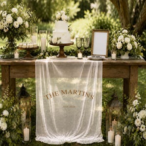 May include: A rustic wooden table draped with a sheer white fabric runner featuring the text THE MARTINS EST. 2026. The table is decorated with a two-tier white cake, white floral arrangements, glass candle holders, and a blank picture frame.