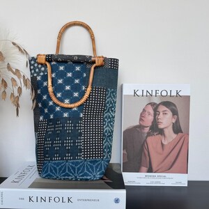 Puede incluir: Un bolso tote vertical confeccionado con tela de patchwork en tonos azul, negro y blanco con varios patrones geométricos. El bolso cuenta con dos asas redondeadas de bambú fijadas en la parte superior. Está junto a una revista Kinfolk.