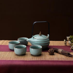 May include: A light blue ceramic tea set consisting of a teapot with a black handle and a tiered, ribbed body, accompanied by four matching small, round tea cups. The pieces feature a subtle crackle glaze finish and brown rim detailing.