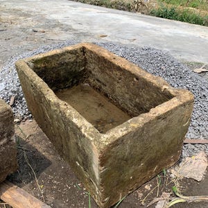 May include: A weathered, rectangular stone trough with mossy green and brown coloration. The trough is filled with a small amount of water and sits on a dirt surface with gravel and a wooden plank nearby. The trough is likely used for gardening or as a water feature.
