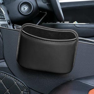 May include: A black car storage pocket made from a leather-like material. The pocket is designed to attach to the car's interior, offering storage for small items. The inside of the pocket is lined with a soft, dark fabric.
