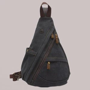 Könnte beinhalten: Anthrazitgraue Canvas-Umhängetasche mit braunen Lederakzenten. Die Tasche verfügt über mehrere Reißverschlussfächer, eine kleine Vordertasche und einen braunen Schultergurt. Die Tasche hat eine dreieckige Form und einen braunen Griff.