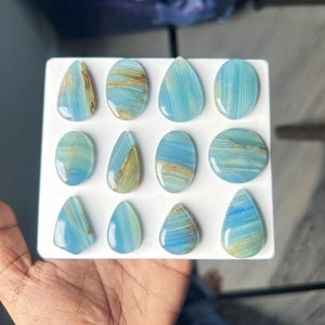 May include: A collection of twelve polished, smooth cabochons arranged in a grid on a white square display board. Each stone features a translucent light blue base color with distinct, horizontal or diagonal golden-brown mineral inclusions.