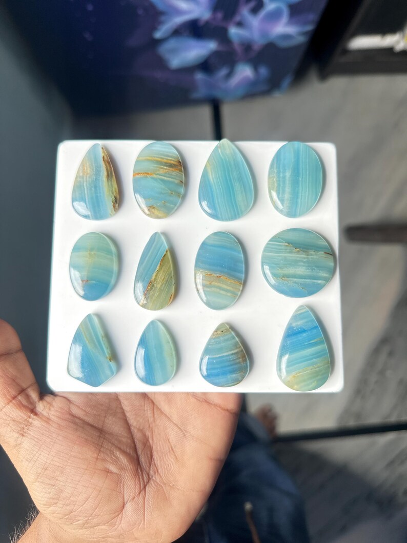 May include: A collection of twelve polished, smooth cabochons arranged in a grid on a white square display board. Each stone features a translucent light blue base color with distinct, horizontal or diagonal golden-brown mineral inclusions.