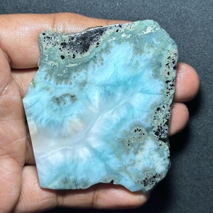 May include: A polished slab of Larimar gemstone featuring a vibrant sky-blue and white marbled pattern with distinct, irregular dark grey and black mineral inclusions along the top and right edges, showcasing a smooth, flat surface texture.