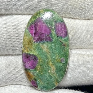 Puede incluir: Gema ovalada con una vibrante mezcla de colores. La piedra presenta una base verde con llamativas manchas de magenta intenso y toques dorados. La superficie pulida refleja la luz, resaltando los patrones naturales.
