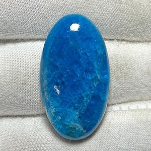 Puede incluir: Una gema ovalada pulida en un tono azul vibrante y saturado. La piedra tiene una superficie texturizada con estructuras cristalinas internas, que reflejan la luz. La gema se encuentra sobre un fondo de tela neutra blanquecina.