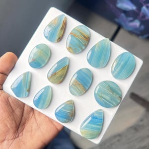 May include: A collection of twelve polished blue onyx cabochons in various shapes, including teardrops, ovals, and circles. Each stone features distinct horizontal banding with shades of light blue, turquoise, and streaks of tan or brown mineral inclusions.