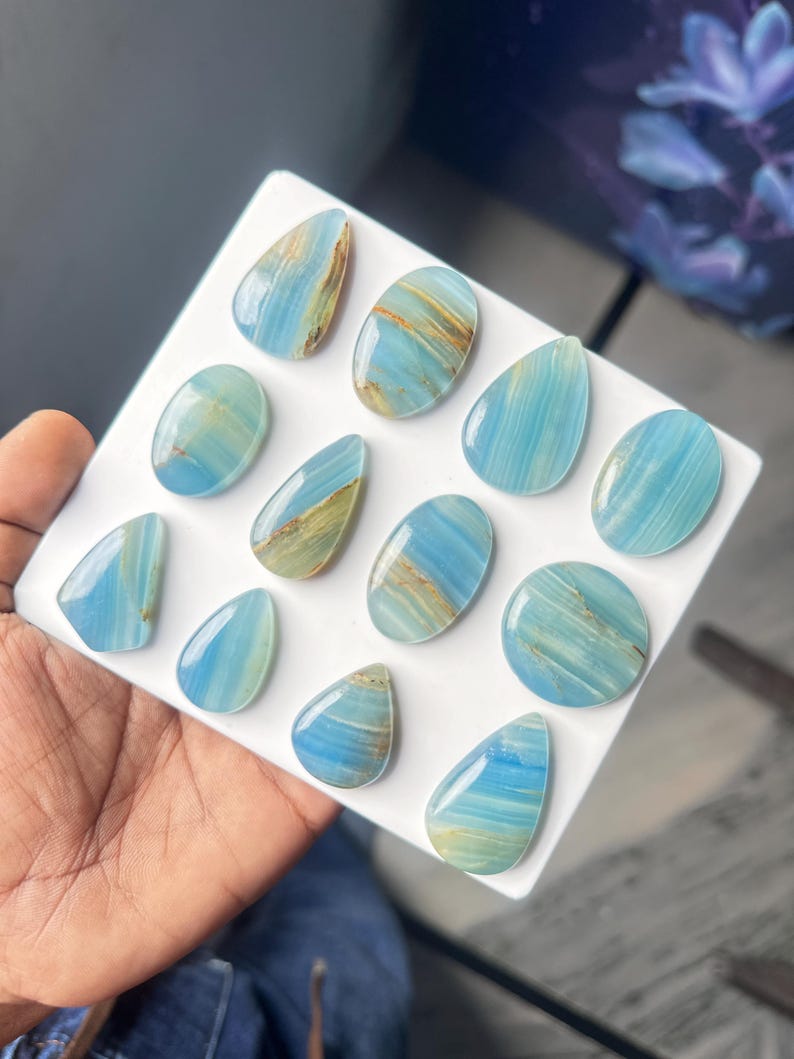 May include: A collection of twelve polished blue onyx cabochons in various shapes, including teardrops, ovals, and circles. Each stone features distinct horizontal banding with shades of light blue, turquoise, and streaks of tan or brown mineral inclusions.