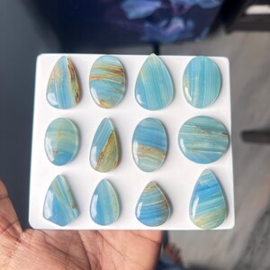 May include: A collection of twelve polished blue gemstones arranged in a grid on a white display board. The stones feature varied shapes including teardrops, ovals, and circles, all displaying light blue hues with distinct brown and tan mineral veining.