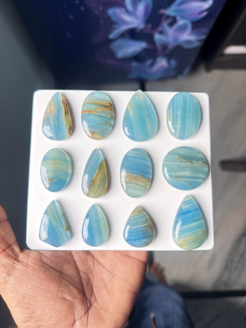 May include: A collection of twelve polished blue gemstones arranged in a grid on a white display board. The stones feature varied shapes including teardrops, ovals, and circles, all displaying light blue hues with distinct brown and tan mineral veining.