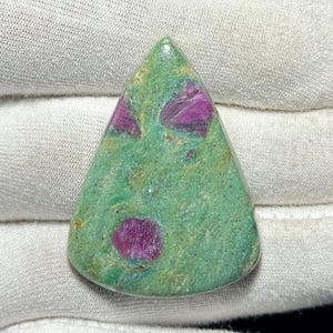 Puede incluir: Una gema triangular con una base verde e inclusiones magenta vibrantes. La piedra pulida tiene una superficie lisa y un patrón único, perfecto para la creación de joyas o coleccionar. Mide aproximadamente 5 cm.