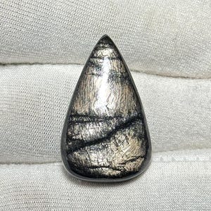 Könnte beinhalten: Ein tropfenförmiger Edelstein mit polierter, glänzender Oberfläche. Der Stein weist eine Mischung aus Silber- und Schwarztönen mit schimmerndem, metallischem Aussehen auf. Der Edelstein ist vor einem weißen Stoffhintergrund platziert.