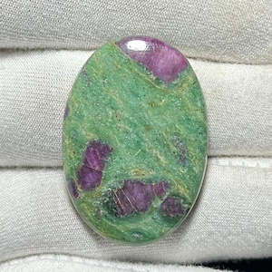 May include: Oval-shaped gemstone with a green base color and vibrant purple inclusions. The polished surface of the stone reflects light, highlighting the natural patterns and textures. This unique gemstone is ideal for jewelry making or collecting.