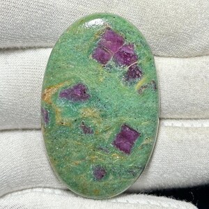 Op de afbeelding: Ovale edelsteen met een groene basiskleur en magenta insluitsels. Het gepolijste oppervlak toont de natuurlijke patronen en texturen van het mineraal. Deze unieke edelsteen is geschikt voor sieraden maken of verzamelen. Afmetingen: circa 3 cm x 2 cm.