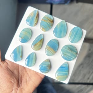 May include: A collection of twelve polished blue gemstones arranged on a white square display board. The stones feature varied shapes including teardrops, ovals, and circles, all displaying light blue hues with subtle golden-brown mineral veining.