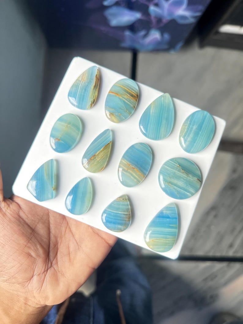May include: A collection of twelve polished blue gemstones arranged on a white square display board. The stones feature varied shapes including teardrops, ovals, and circles, all displaying light blue hues with subtle golden-brown mineral veining.