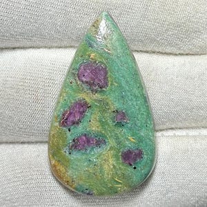 Puede incluir: Gema en forma de lágrima con base verde y inclusiones rojo-púrpura. La piedra pulida tiene un aspecto natural. La gema es adecuada para la fabricación de joyas o coleccionismo. Mide aproximadamente 4 cm de largo.